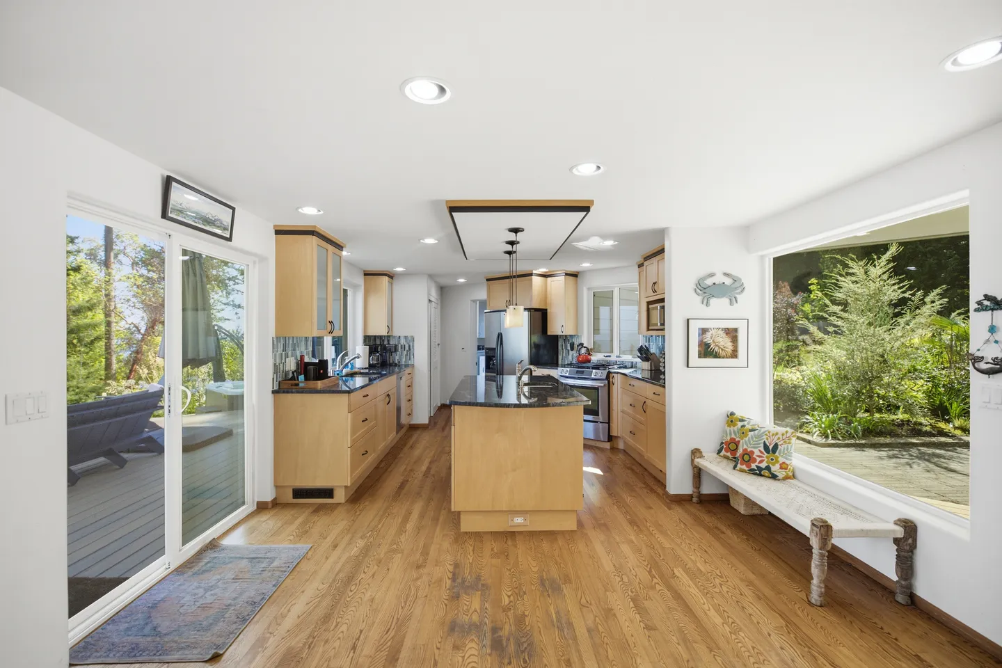 Open-concept kitchen with light wood cabinets, dark countertops, a central island, and hardwood floors next to sliding glass doors and a large picture window.