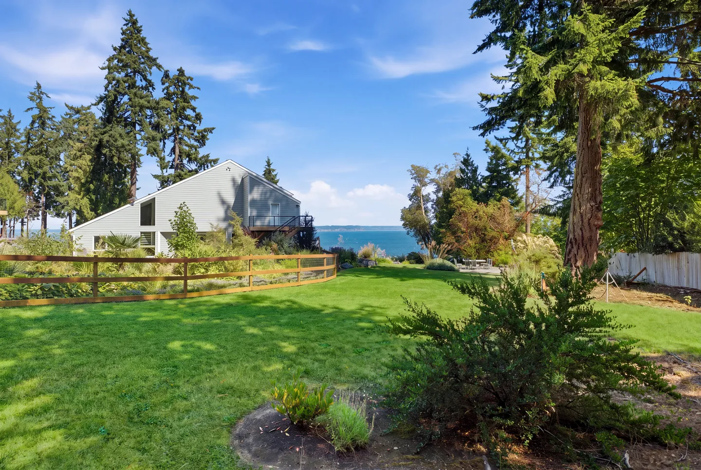 Spacious green lawn behind a wooden split-rail fence leading toward a grey two-story house with a water view in the distance.