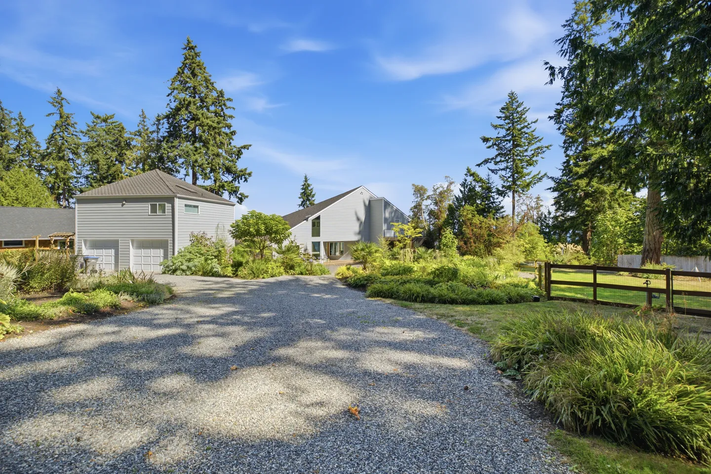 A gravel driveway leading up to a two-story garage and a contemporary house surrounded by lush greenery under a clear blue sky.