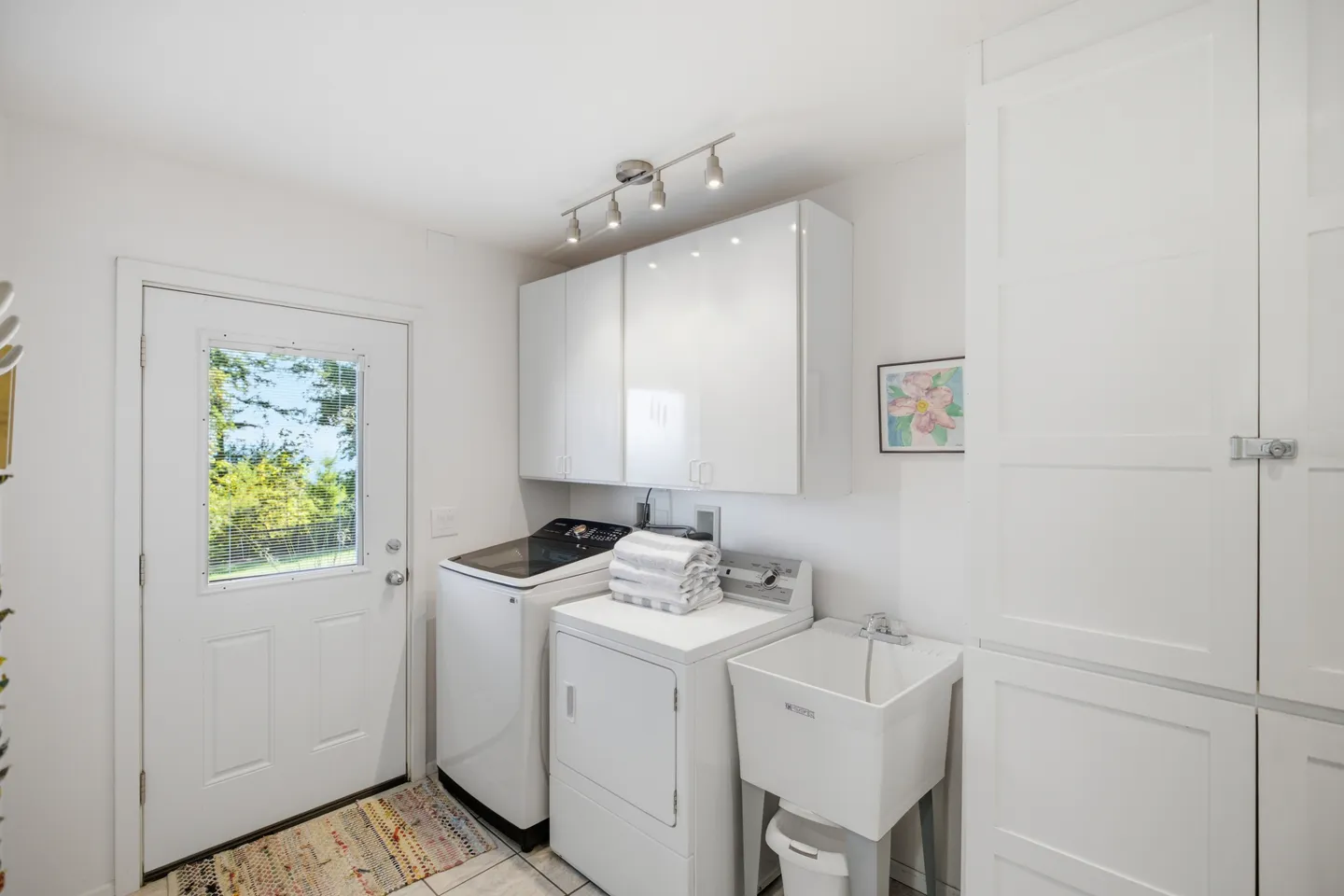 A laundry room with a white washing machine, dryer, utility sink, white cabinets, and an exterior door with a window.