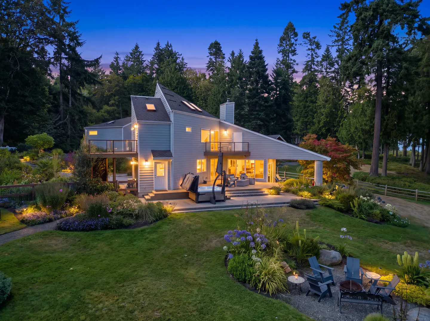 Dusk view of the exterior back of a multi-level house with a patio, hot tub, fire pit seating area, and lush green lawn surrounded by tall evergreen trees.