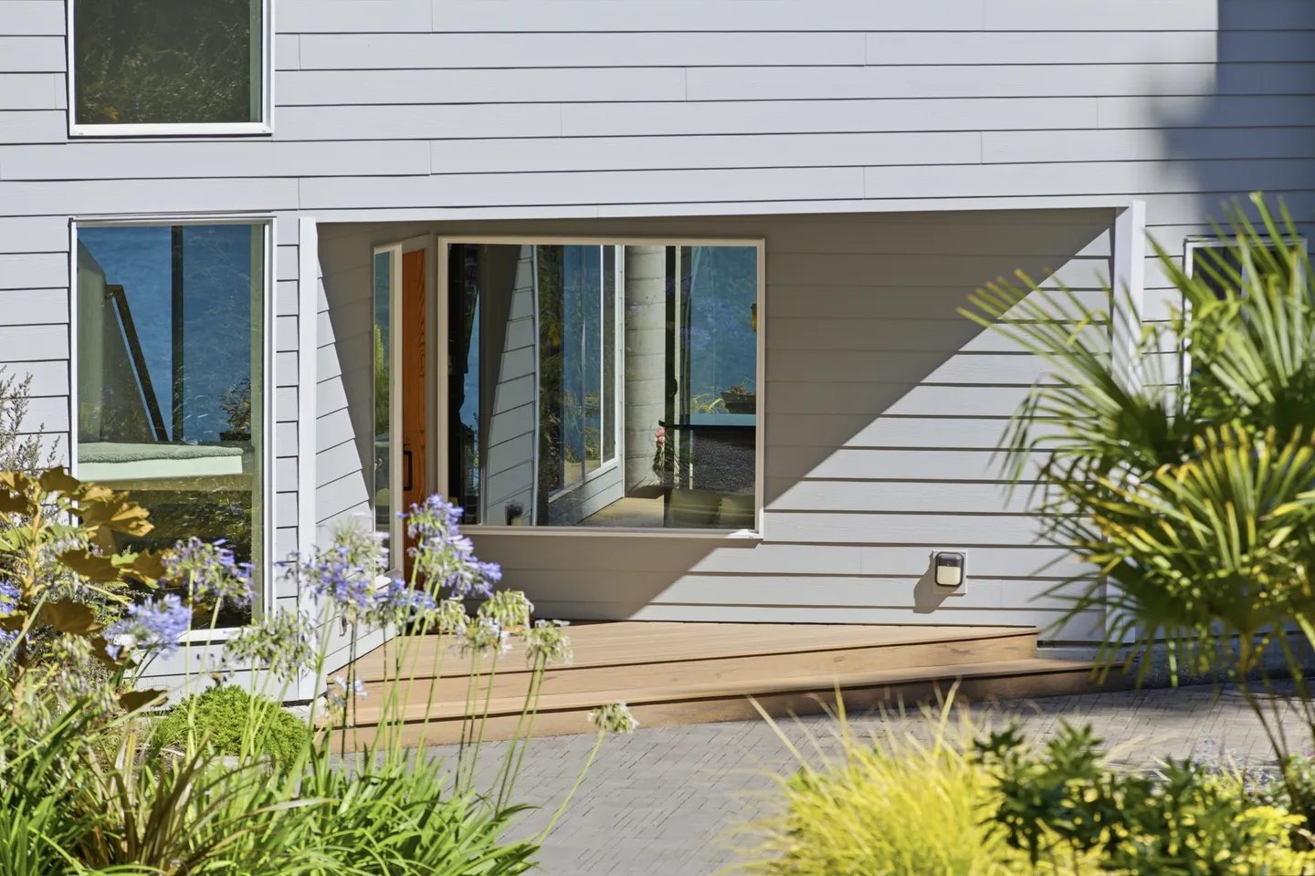 Exterior view of a home entrance with horizontal siding, wooden steps, and large glass windows surrounded by landscaping.