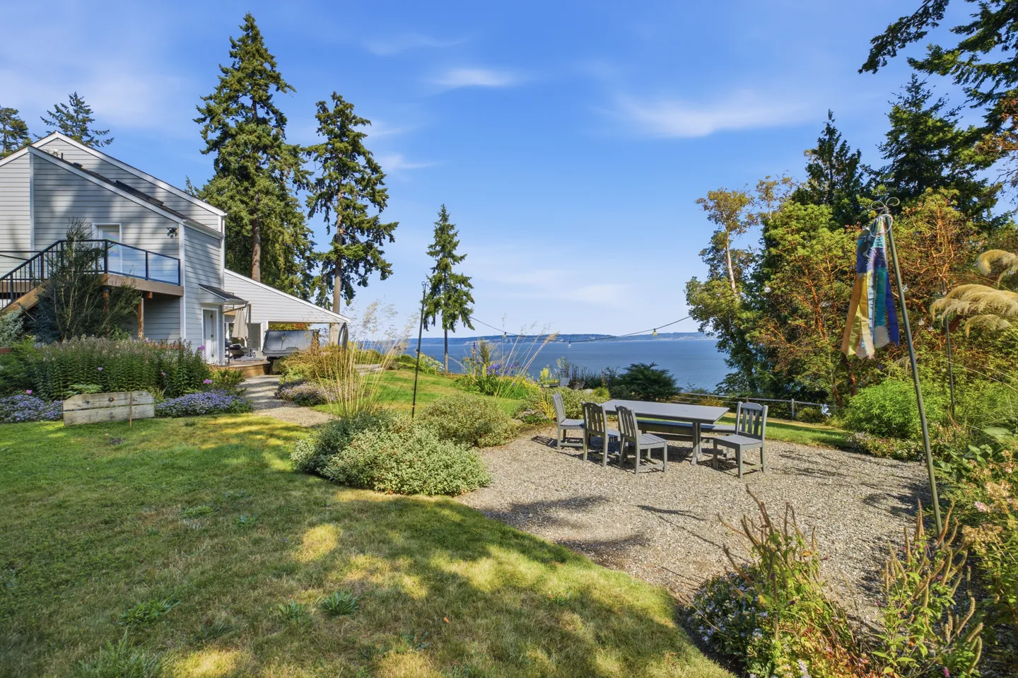 A backyard with a gravel dining area, outdoor table and chairs, overlooking a wide body of water surrounded by trees.