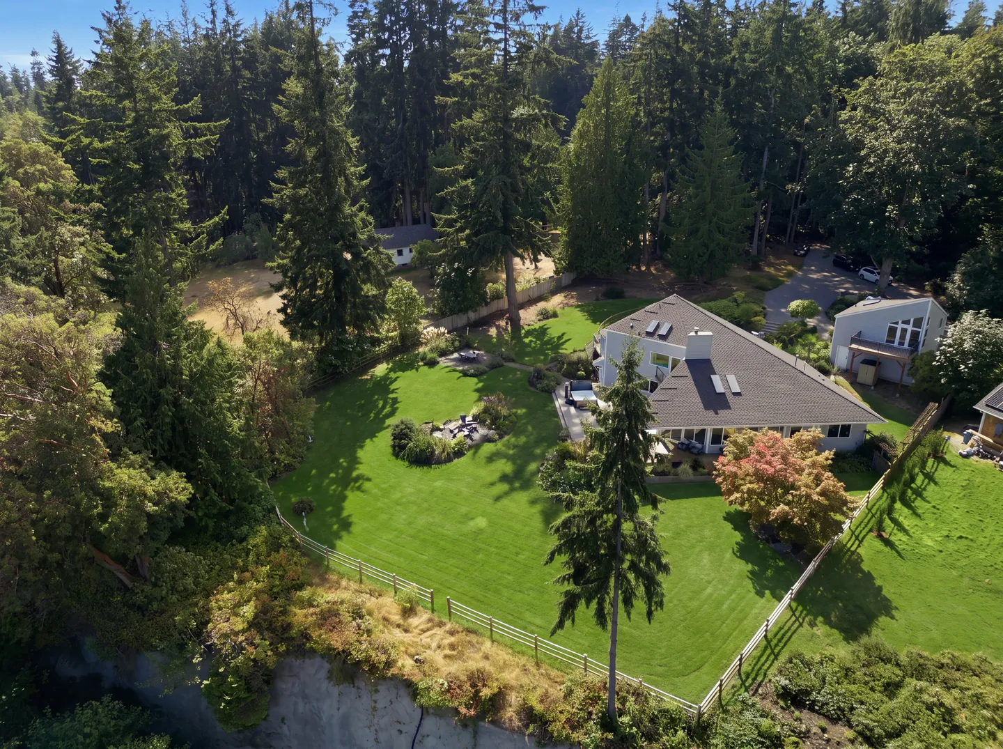 Aerial view of a house with a fenced grassy lawn surrounded by evergreen trees near a cliff edge.