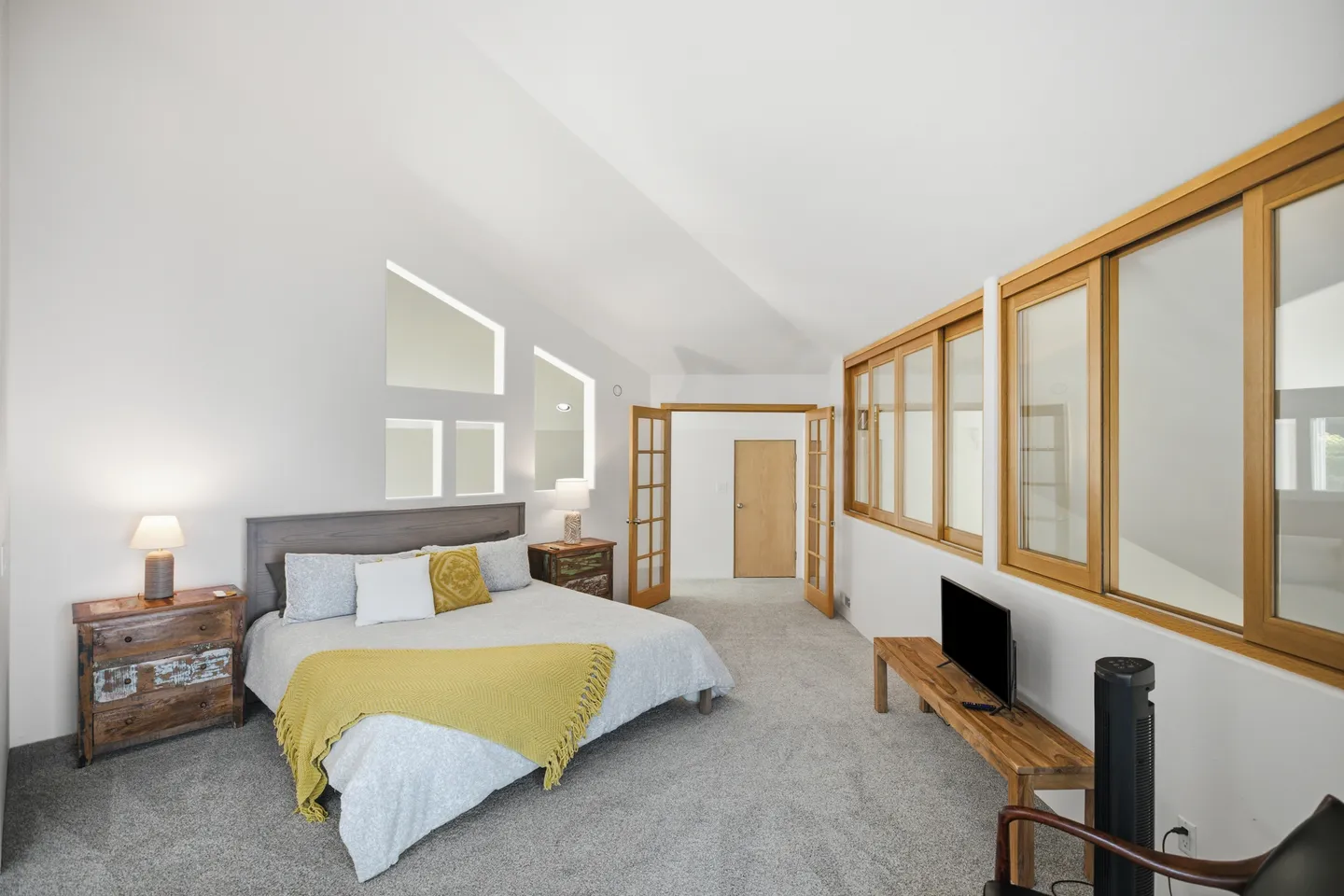 Spacious bedroom with vaulted ceilings, carpeted flooring, a bed, nightstands, and interior windows with wood frames.