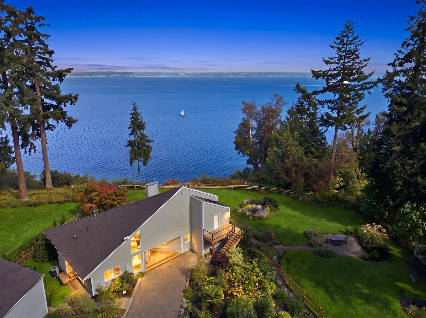 Aerial view of a coastal house surrounded by green lawns and tall evergreen trees, overlooking a large body of water.