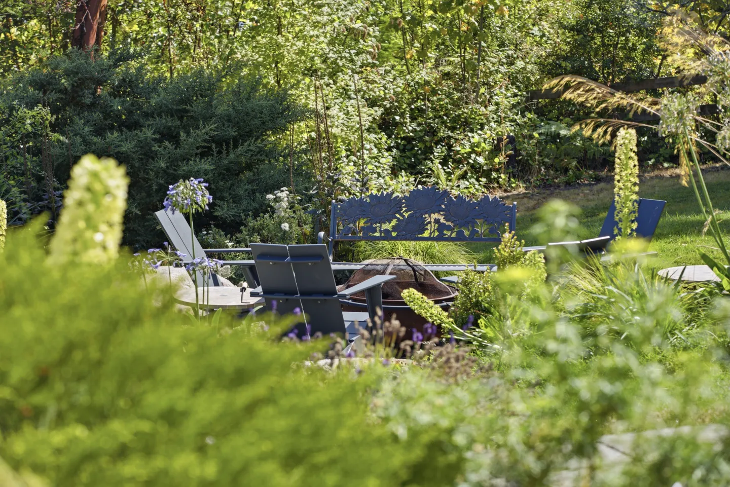 Outdoor garden seating area with a decorative bench, chairs, and a fire pit surrounded by lush greenery.