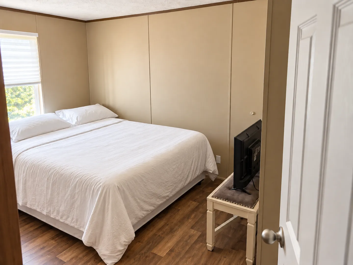 Bedroom with a bed dressed in white linens, a window, wood-look flooring, and a flat-screen television on a small table.