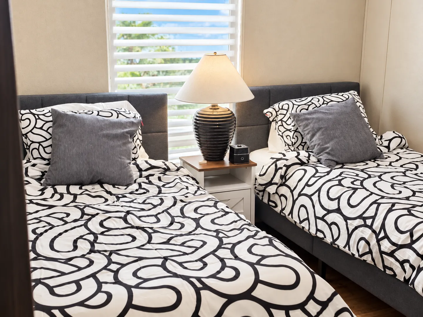 Bedroom with two twin beds dressed in black and white patterned bedding separated by a nightstand and lamp beneath a window.