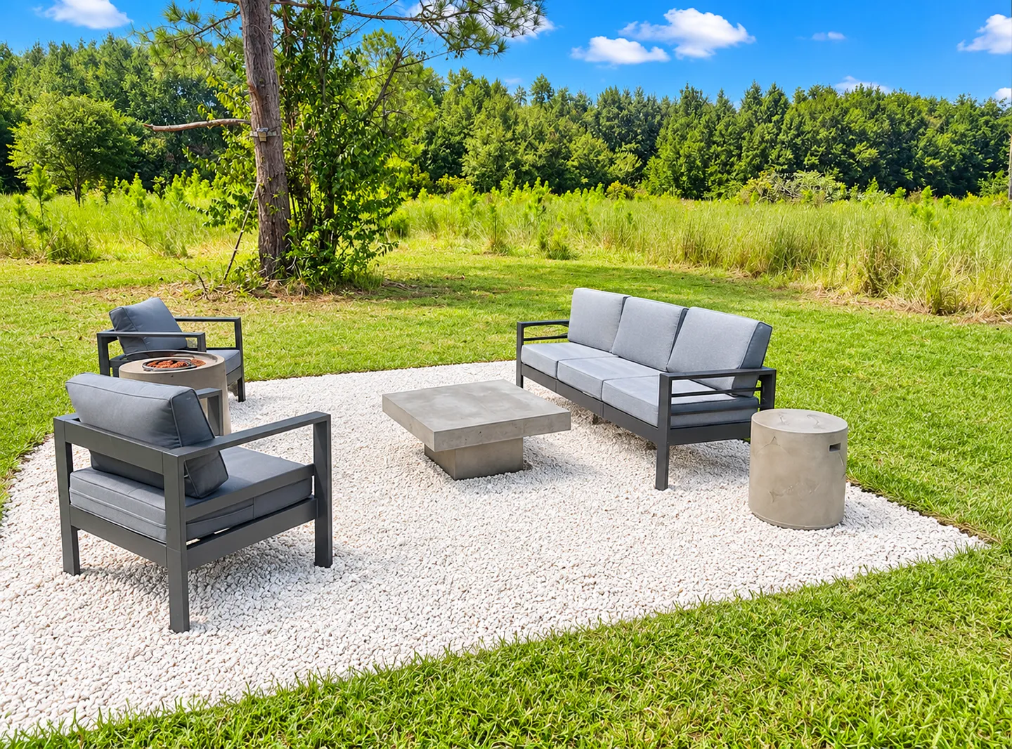 Outdoor gravel patio area with a sofa, two armchairs, a coffee table, and a small fire pit surrounded by green lawn and trees.