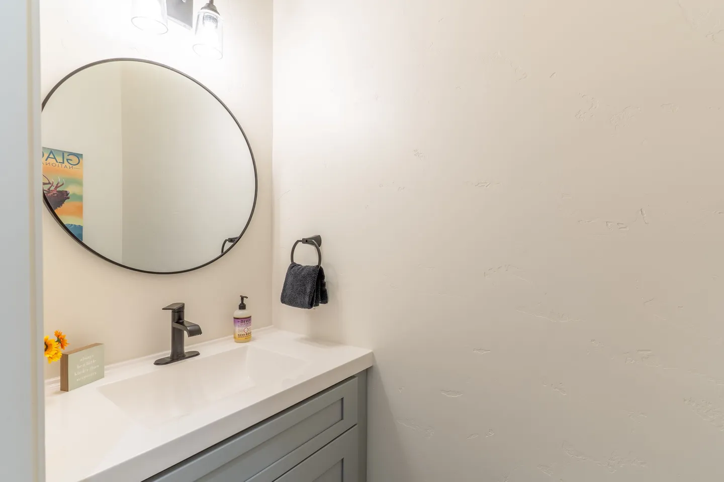 Bathroom vanity with a white countertop, matte black faucet, and a round mirror with a thin black frame on a textured off-white wall.
