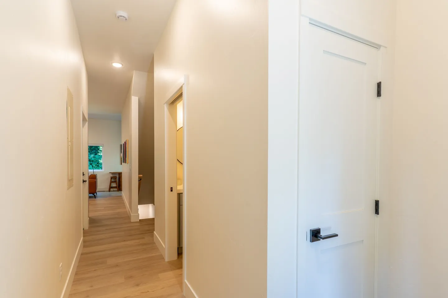 A brightly lit hallway with light wood flooring, neutral walls, white interior doors, and a glimpse into an adjacent bathroom and living area.