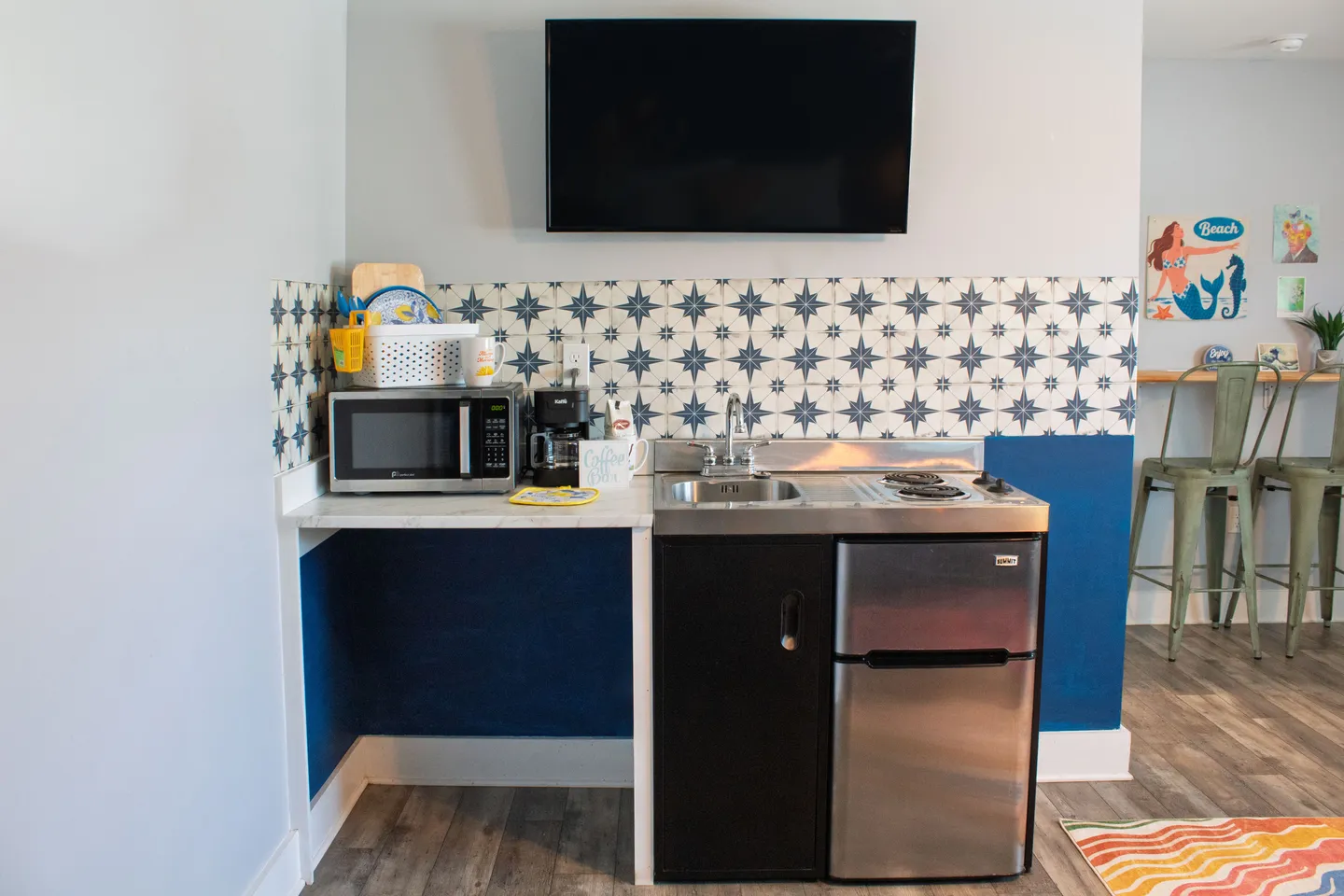 Compact kitchenette featuring a mini fridge, two-burner cooktop, sink, microwave, and coffee maker beneath a wall-mounted TV.