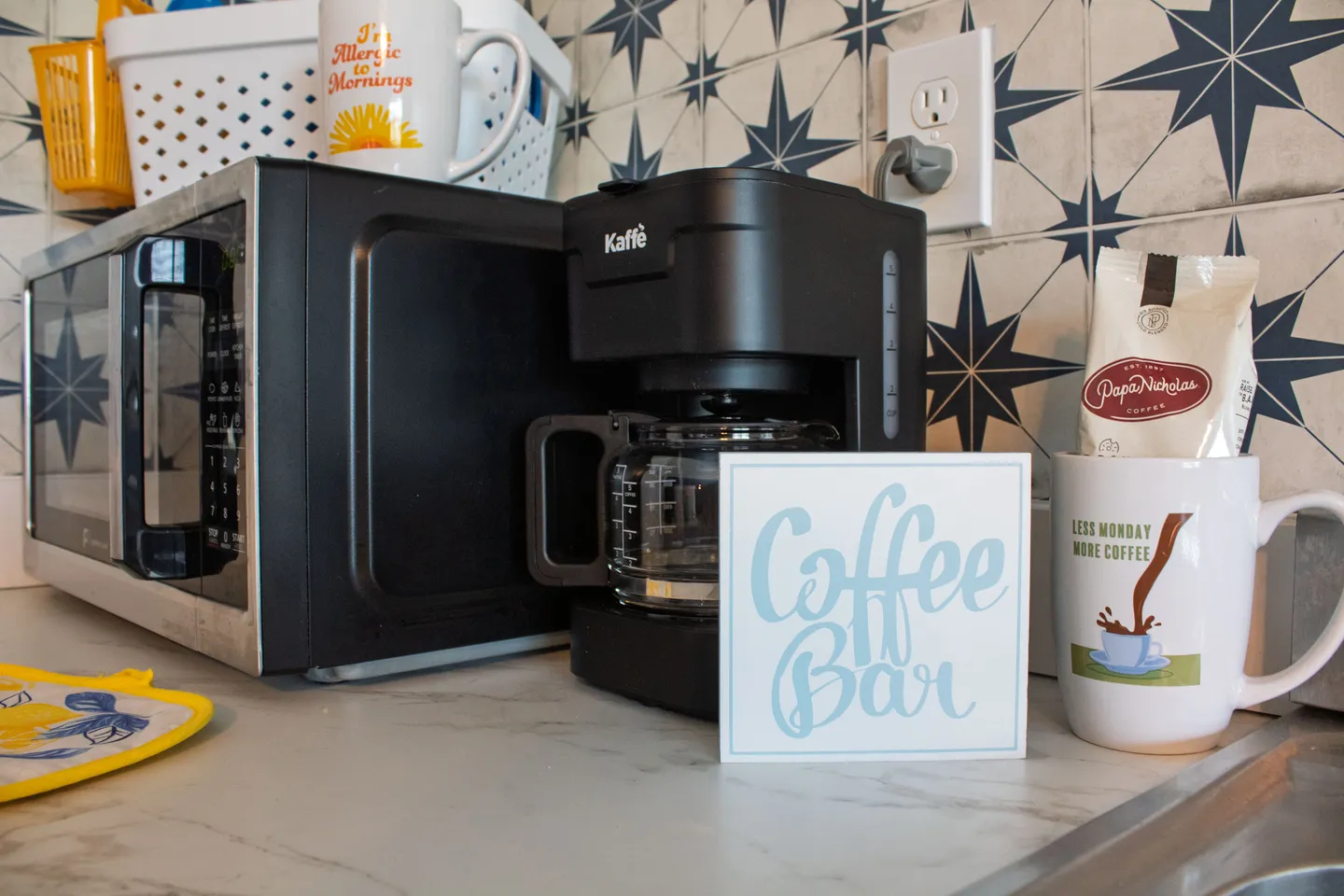 Kitchen counter coffee station with a drip coffee maker, coffee bar sign, microwave, mug, and bag of coffee against a blue patterned tile backsplash.