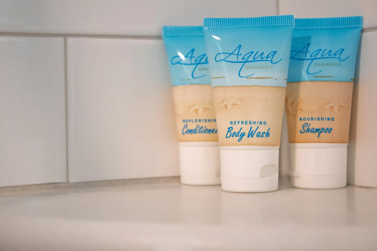 Close-up of travel-sized Aqua Organics shampoo, conditioner, and body wash bottles in a tiled shower or bathroom shelf.