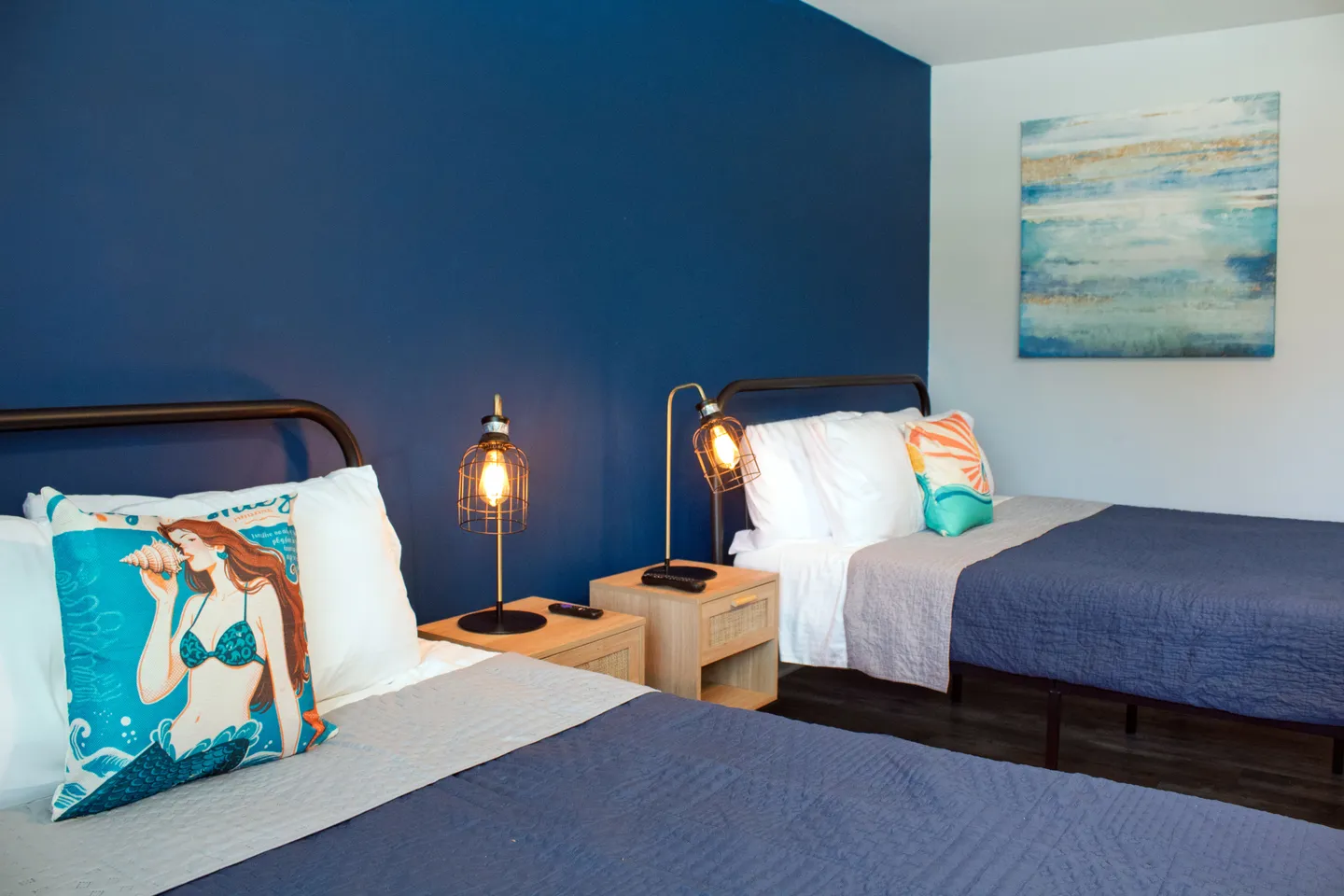 Bedroom with two beds featuring metal headboards, blue bedding, and a shared wooden nightstand against a deep blue accent wall.