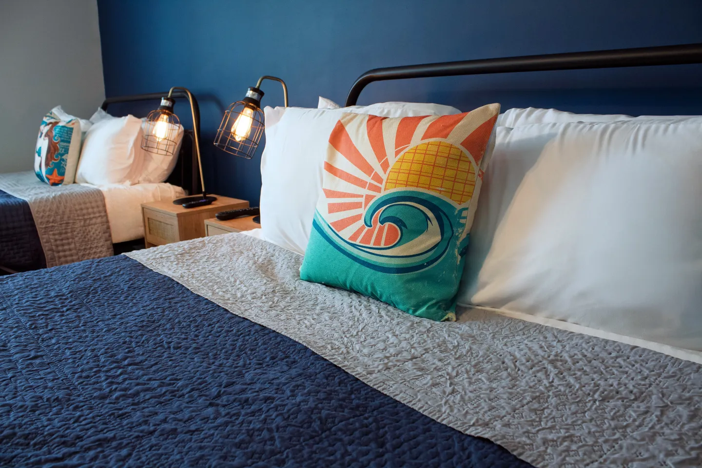 Close-up of a bed with a colorful wave accent pillow in a bedroom with blue walls and a second bed visible nearby.