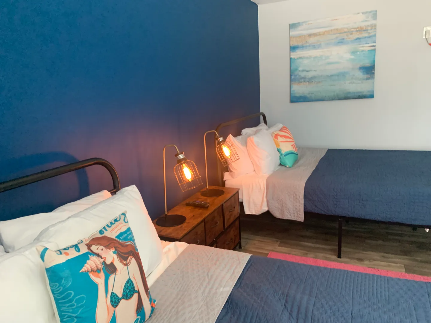 Bedroom with two beds, a blue accent wall, a shared nightstand with industrial lamps, and abstract wall art.