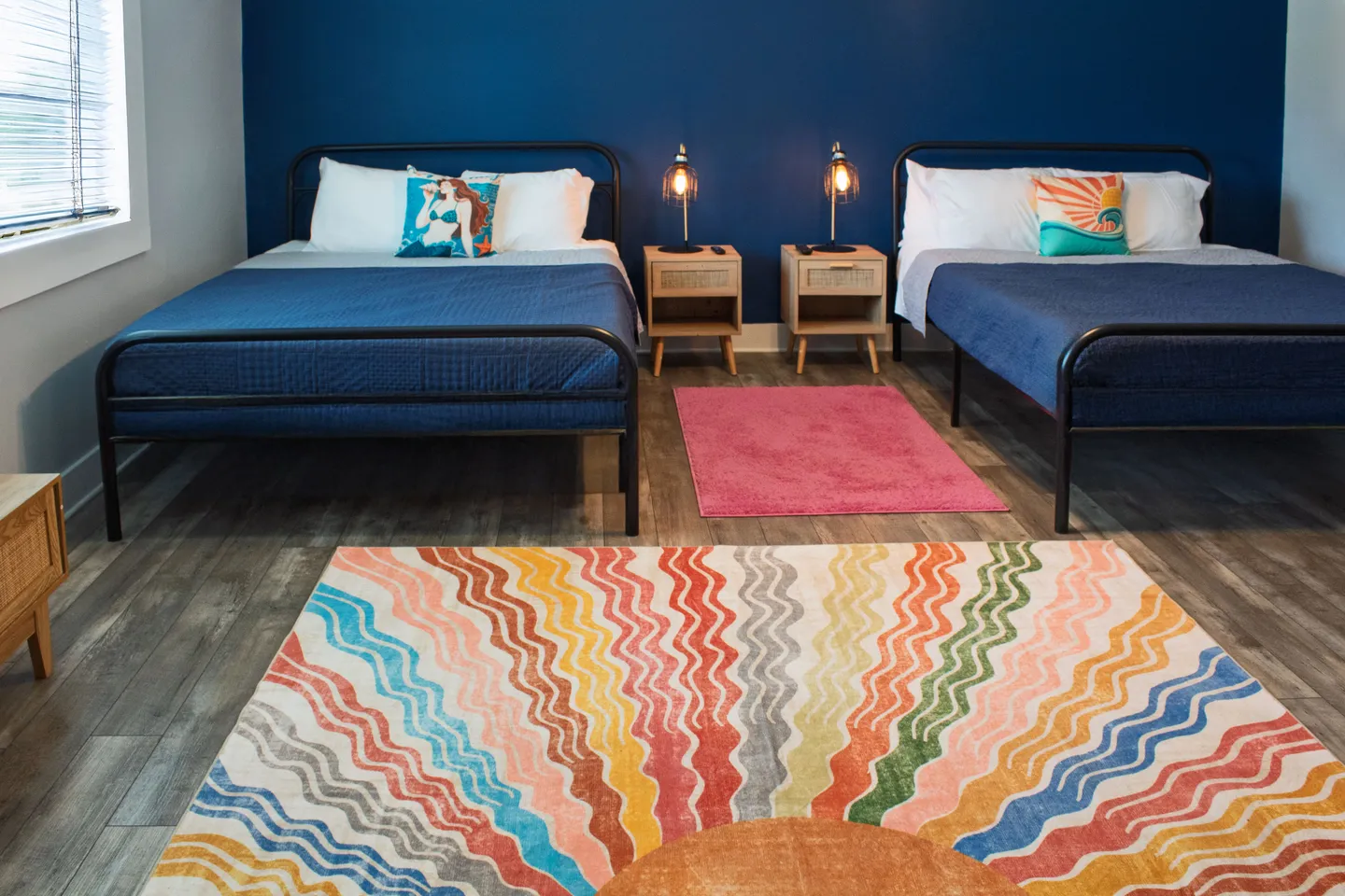 Bedroom with two metal-frame beds, navy blue bedding, a deep blue accent wall, and colorful patterned rugs.