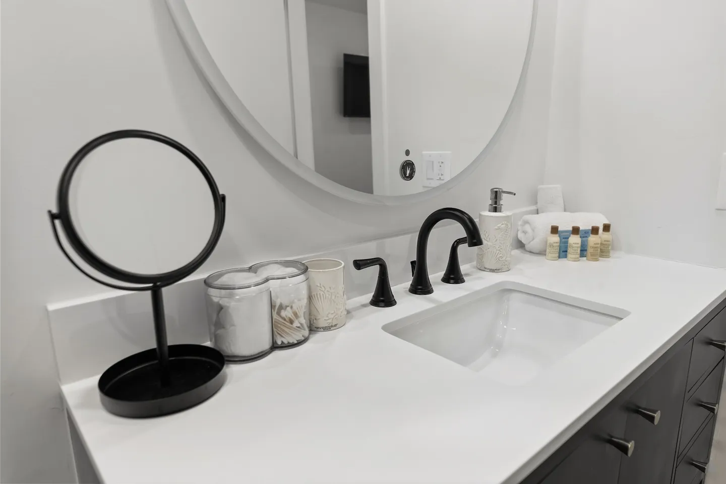 Bathroom vanity with a white countertop, undermount sink, dark fixtures, round mirror, and complimentary toiletries.