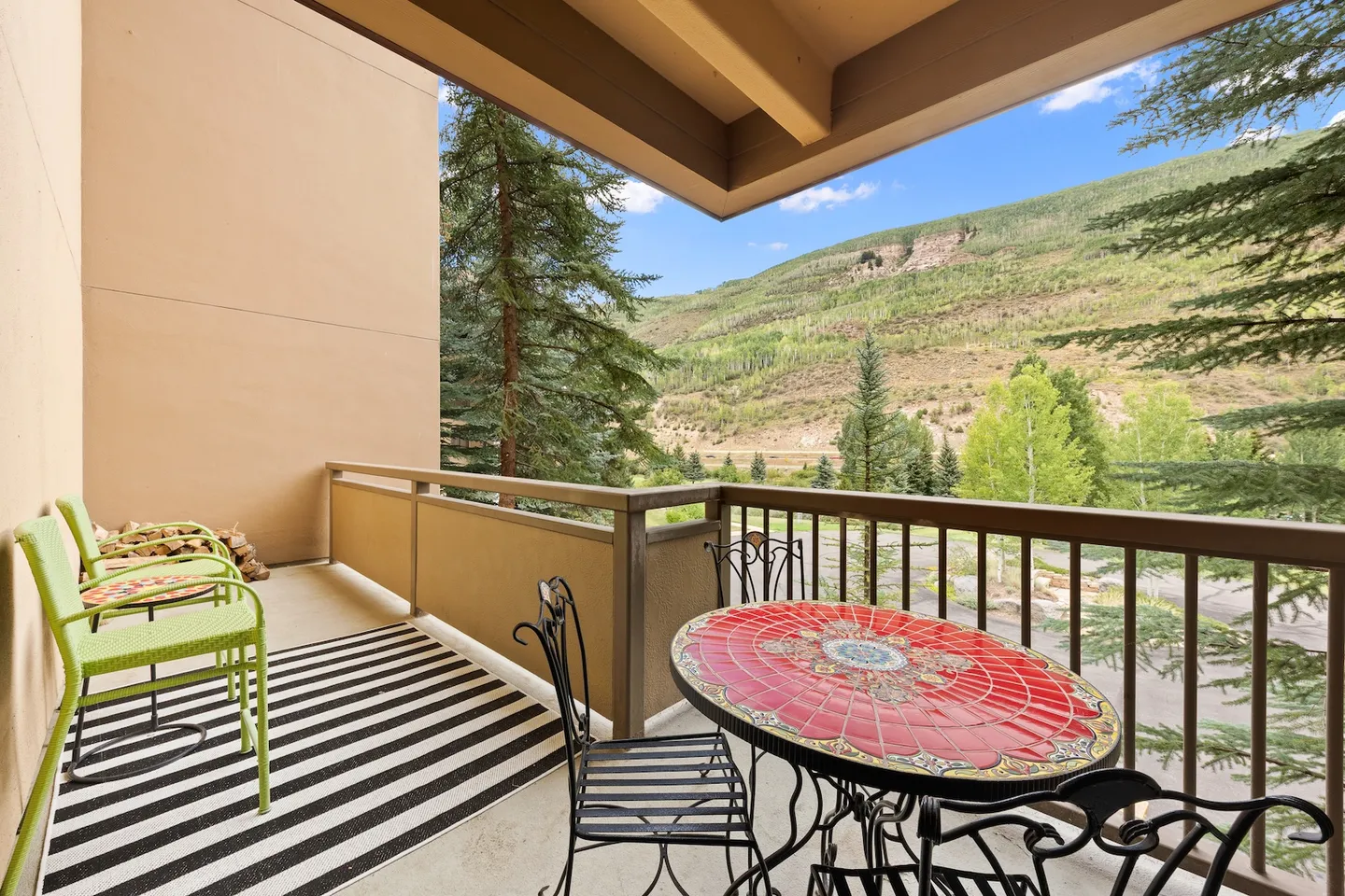 Covered outdoor balcony with a round red mosaic table, wrought-iron chairs, green chairs, and a scenic mountain view.