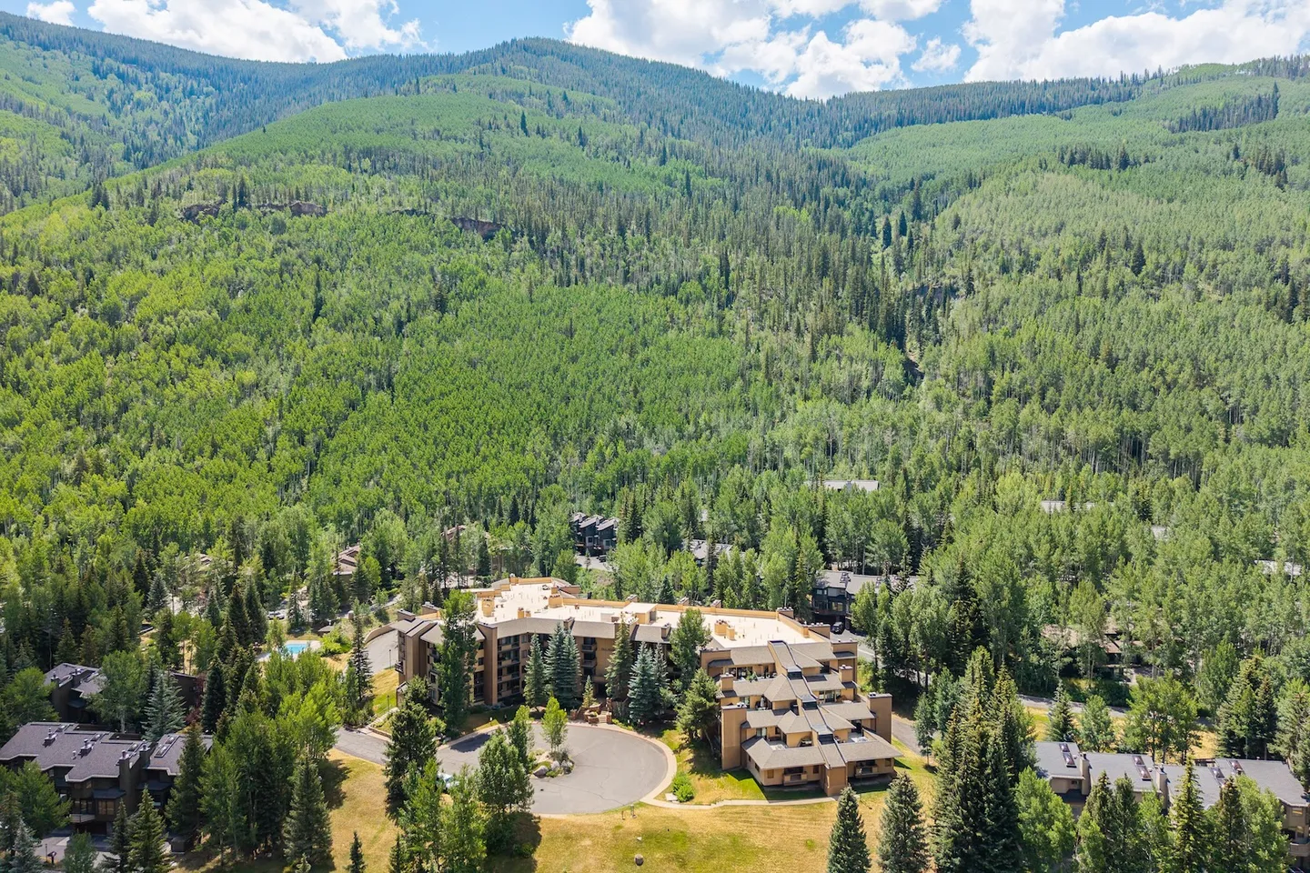 Aerial view of a multi-story lodge and residential complex nestled at the base of a forested mountain on a sunny day.