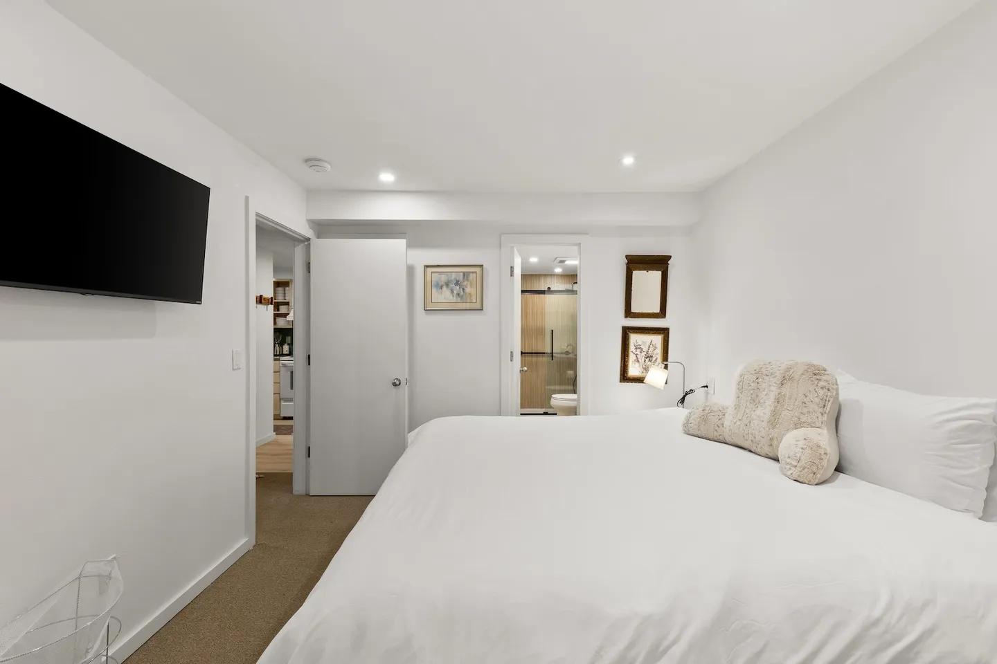 Bedroom with a bed made with white linens, a wall-mounted flat-screen TV, carpeted floors, and views into an ensuite bathroom and hallway.