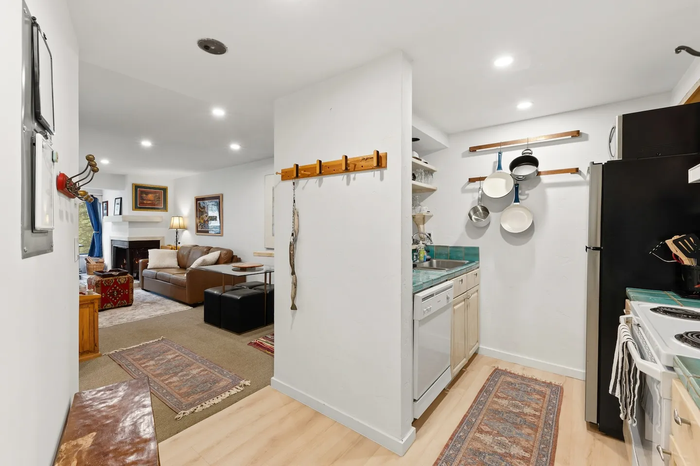 Open view showing a kitchen on the right with hanging cookware and a living room on the left with a brown leather sofa and fireplace.