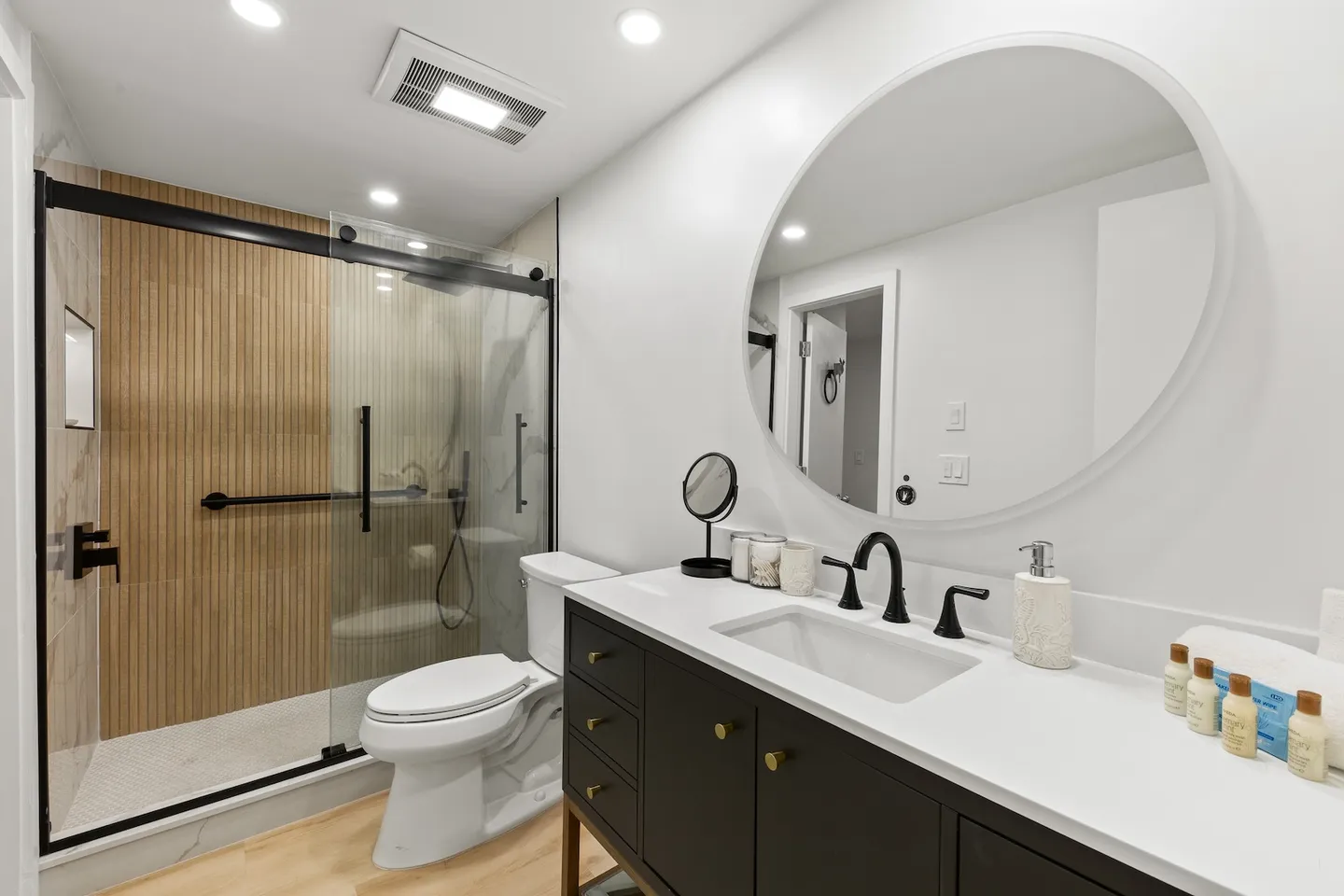Modern bathroom featuring a dark vanity with white countertop, round mirror, toilet, and a glass-enclosed walk-in shower with vertical wood-look tile accents.