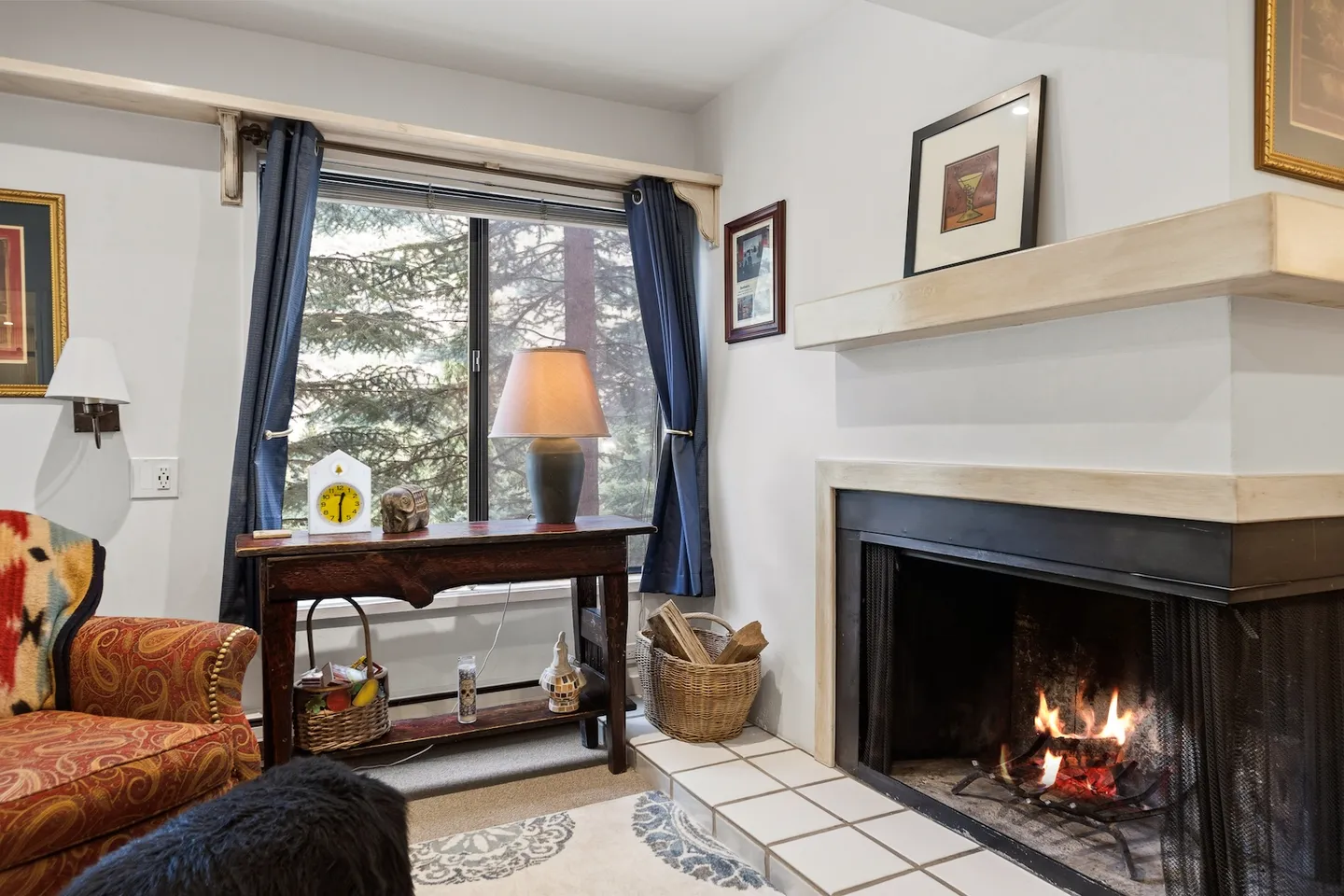 A corner fireplace with a fire burning, adjacent to a wooden table and a window looking out onto trees.