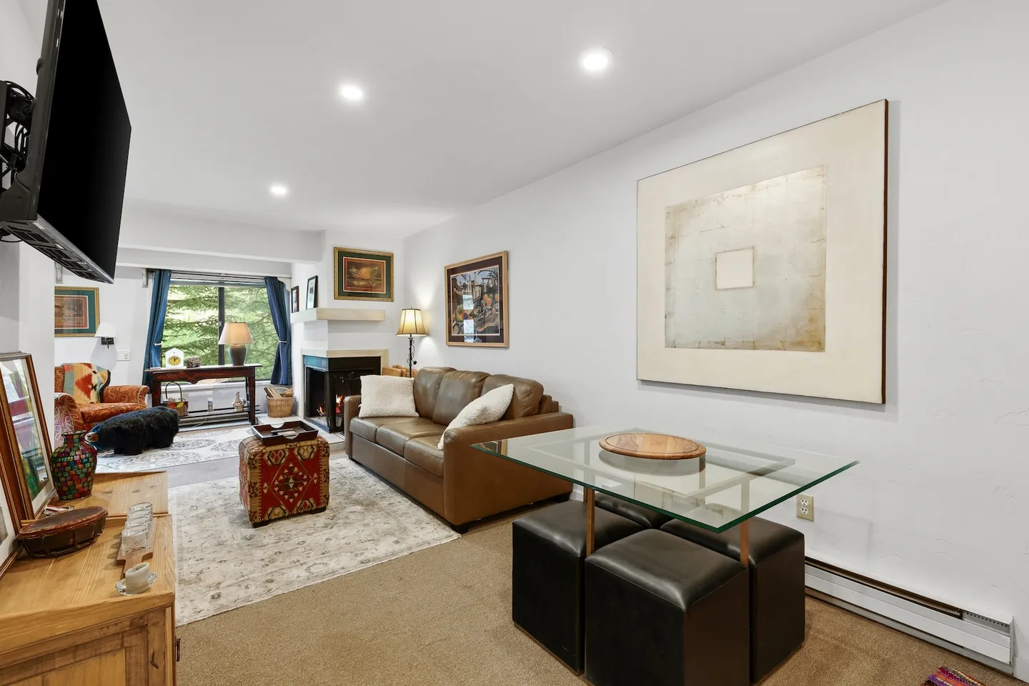 Open living area with a brown leather sofa, patterned rug, glass-topped table with ottomans, and a fireplace in the background.