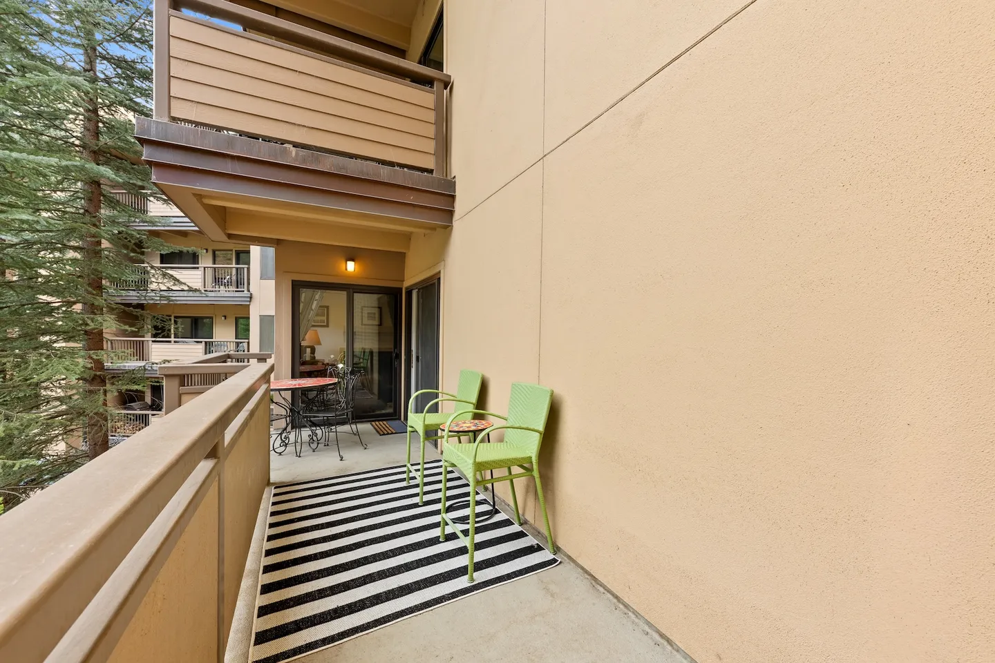 Covered outdoor balcony with lime green chairs, a striped rug, and a small bistro set overlooking trees.