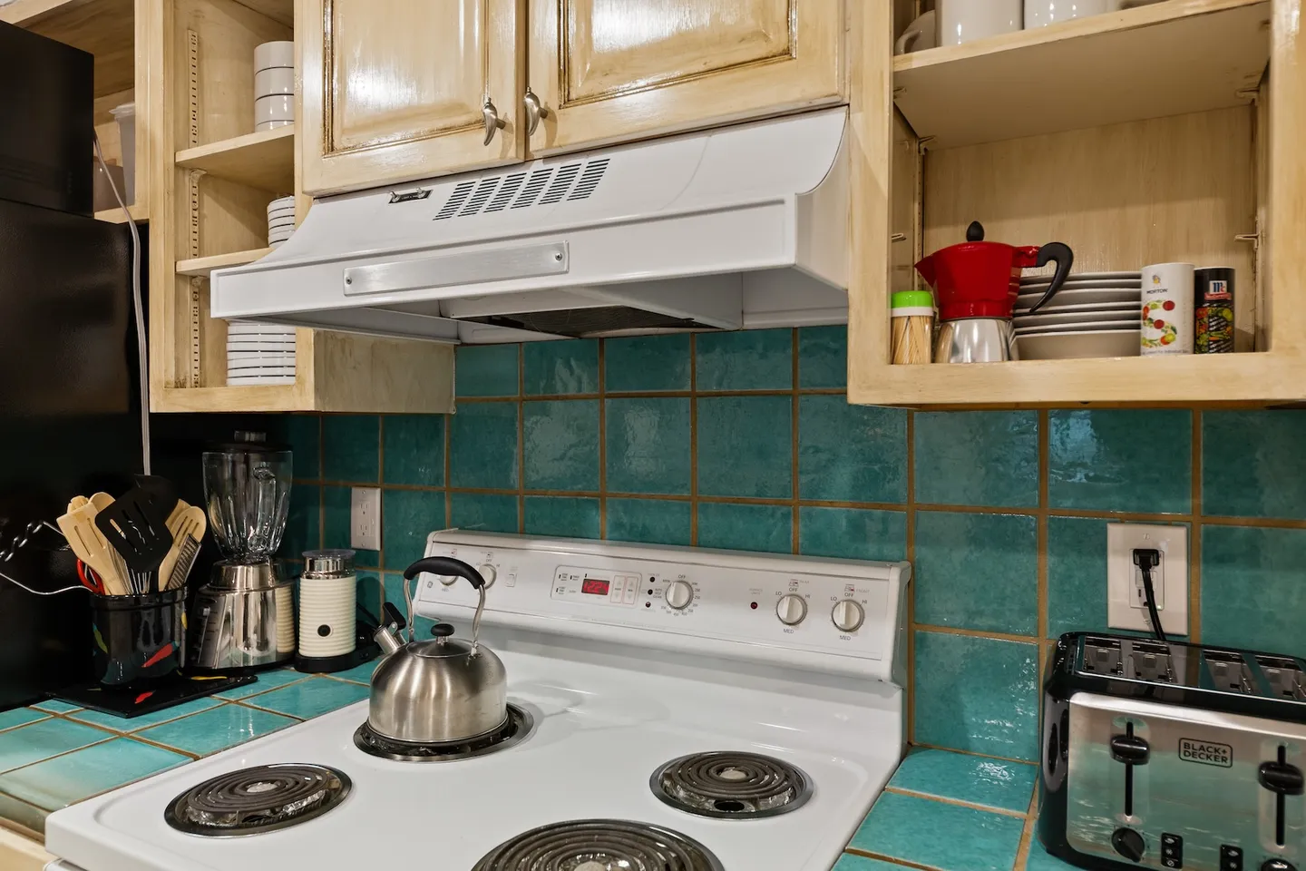 Kitchen counter area with an electric stovetop, tea kettle, teal tile backsplash, and light wood upper cabinets.