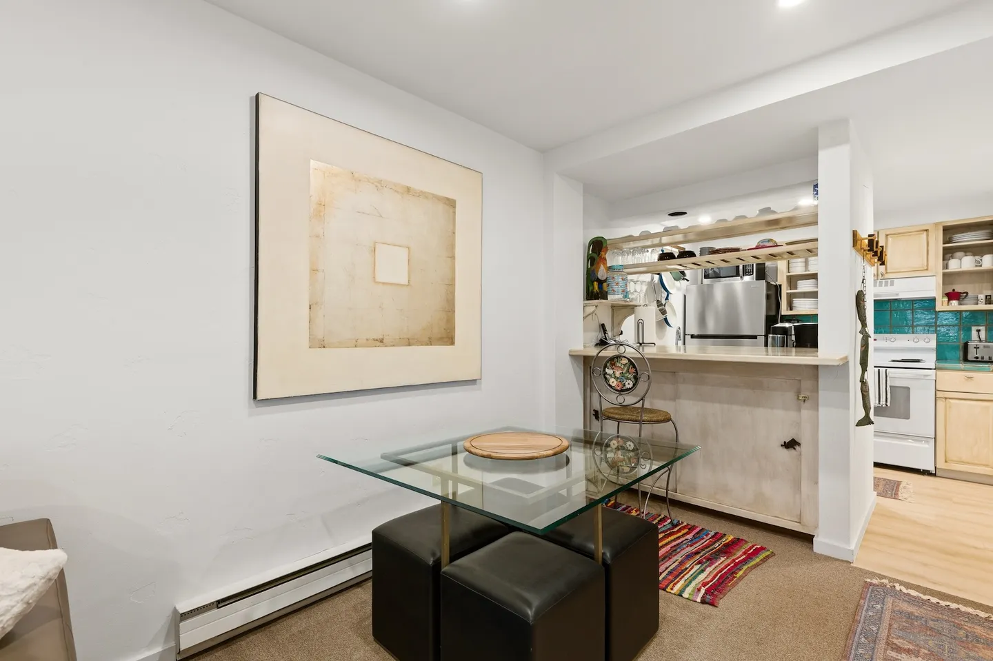 Dining area featuring a glass-top table with dark cube ottomans, large wall art, and an opening with a counter looking into the kitchen.
