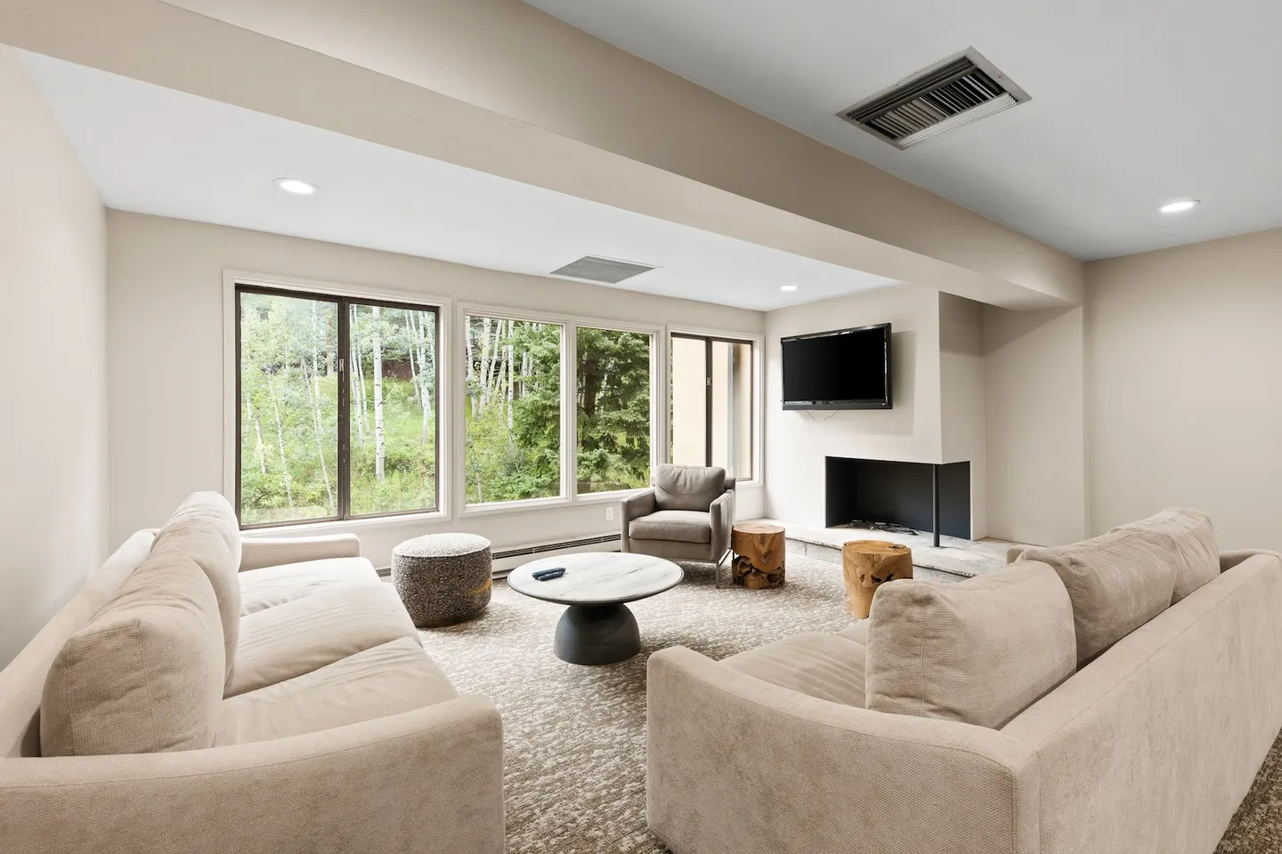 Living room with two neutral fabric sofas, an armchair, a central coffee table, a fireplace, and large windows looking out to trees.