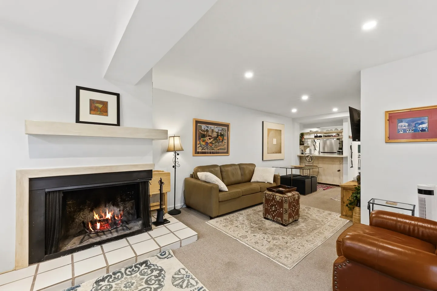 Living room with a lit fireplace, tiled hearth, leather seating, area rug, and an adjoining kitchen nook.