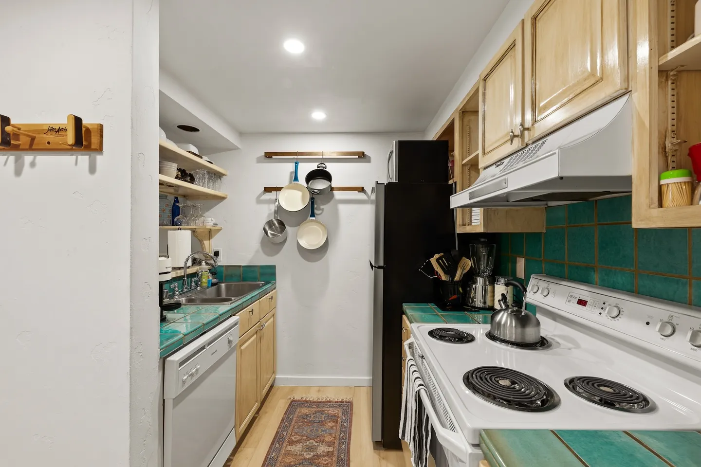 A galley kitchen with turquoise tile countertops, light wood cabinets, white appliances, and hanging pots and pans on the back wall.