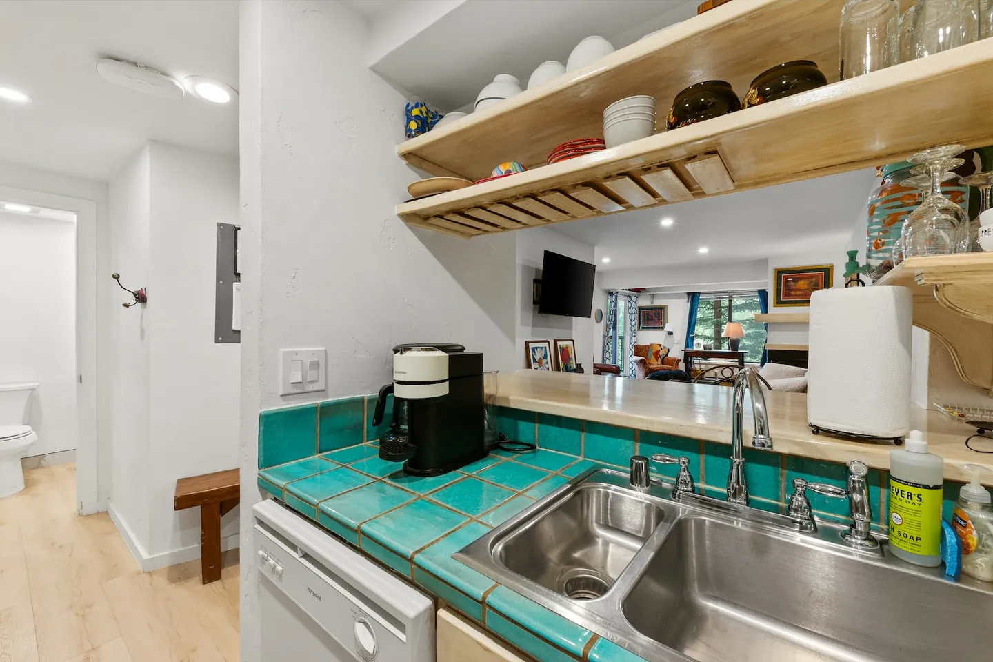 Kitchen counter with teal tile, stainless steel double sink, open wooden shelving, and a view into the living room.