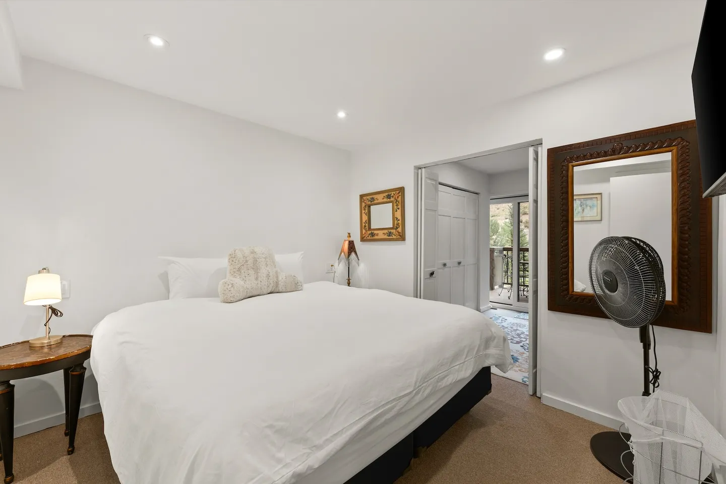 Bedroom featuring a made bed with white linens, a bedside table with a lamp, a large wall mirror, and a doorway leading to another area.