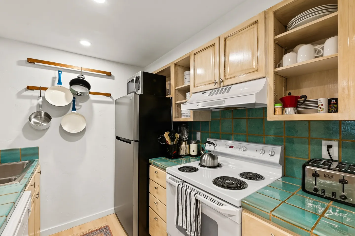 Kitchen with green tile countertops, light wood cabinets, electric white stove, and cookware hanging on the wall.