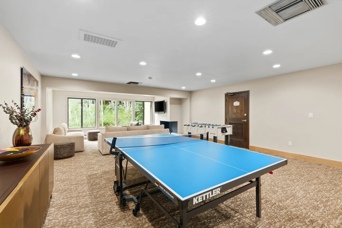 Game room featuring a blue ping pong table, foosball table, and living area with sofas in front of large windows.