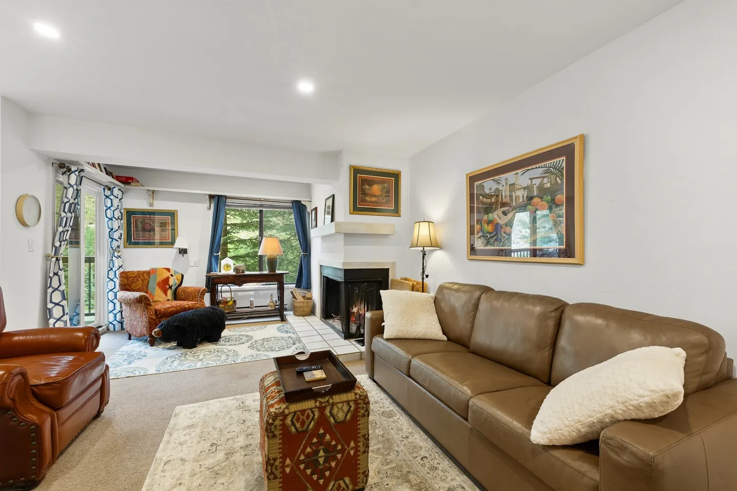 Living room with a brown leather sofa, patterned armchair, wood-burning fireplace, and windows looking out to trees.