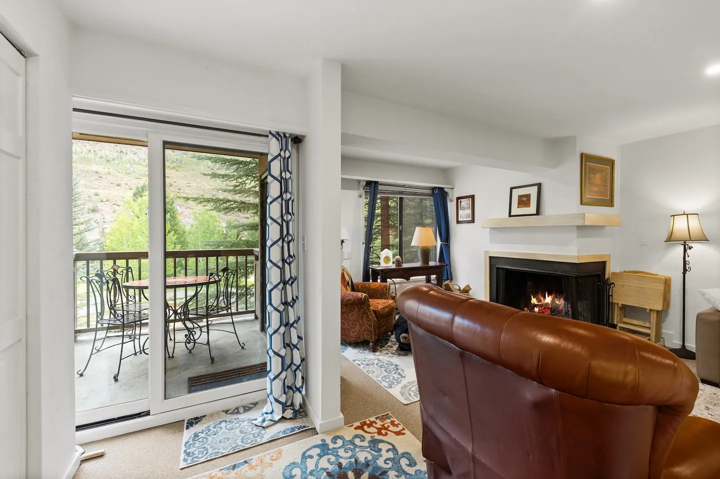 Living room with sliding glass door leading to a balcony, featuring a lit fireplace and a brown leather armchair.