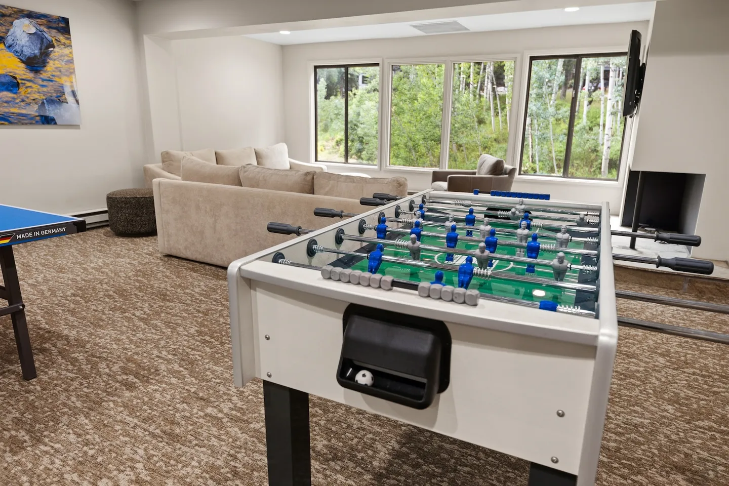 Game room and lounge area with a foosball table, a ping-pong table edge, a sectional sofa, and windows overlooking trees.