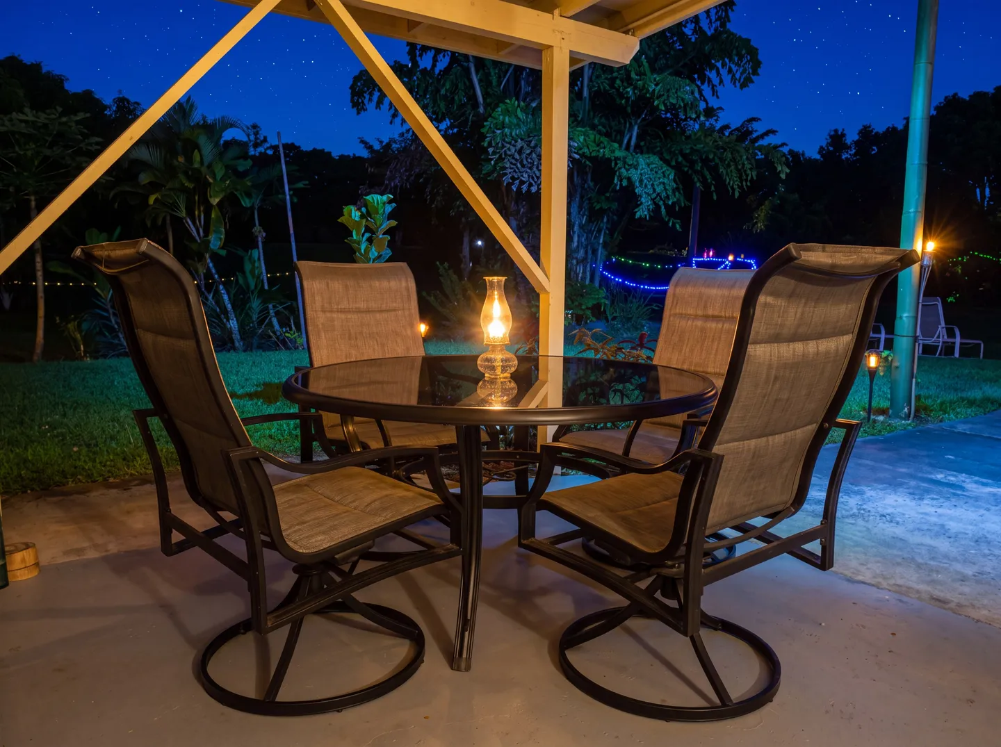 A covered patio at night with a round glass-top dining table, four swivel armchairs, and a lit lantern on the table overlooking a lawn.