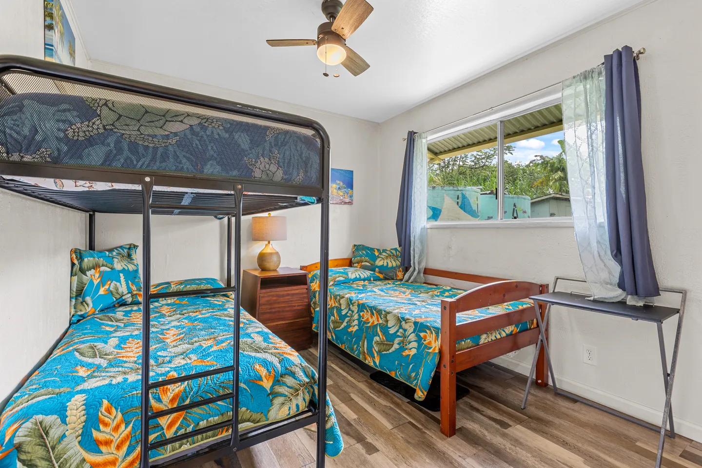 Bedroom with a metal bunk bed, a single wooden bed, a nightstand with a lamp, and a small folding table near a window.