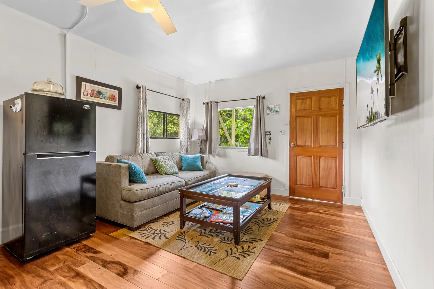 Living area with wood-look flooring, a sofa with throw pillows, a coffee table on an area rug, a compact refrigerator, and a wall-mounted TV.