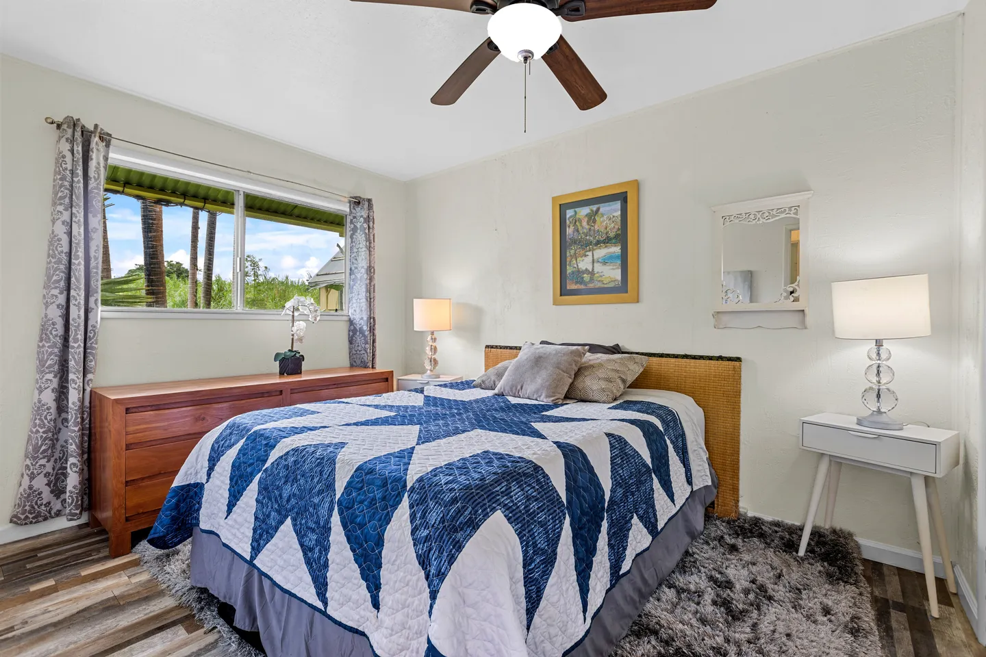 Bedroom with a patterned blue and white quilt on the bed, wooden dresser, nightstands with lamps, and a ceiling fan.