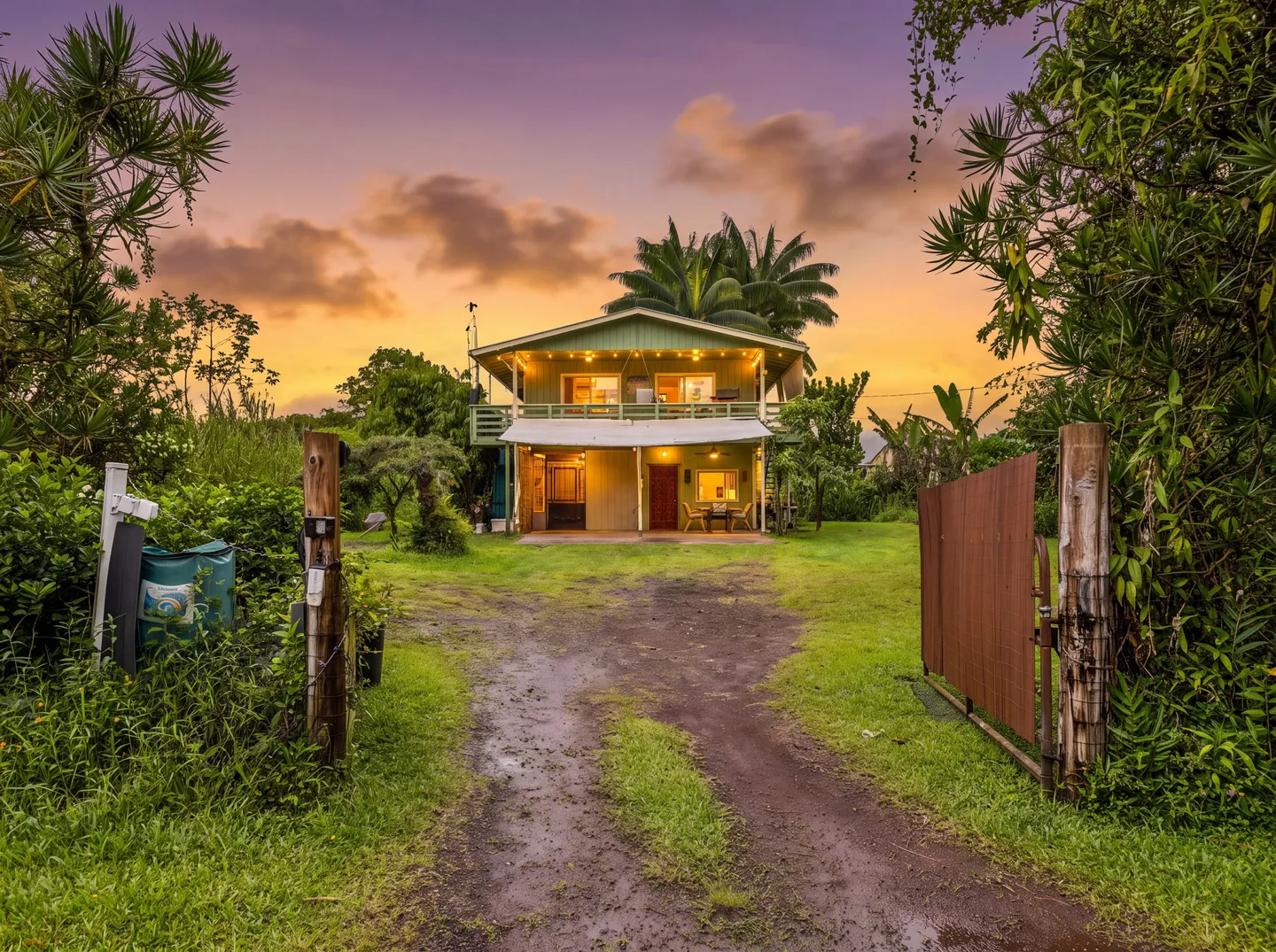 Exterior of a two-story green house with a balcony and patio, viewed down a dirt driveway at sunset.