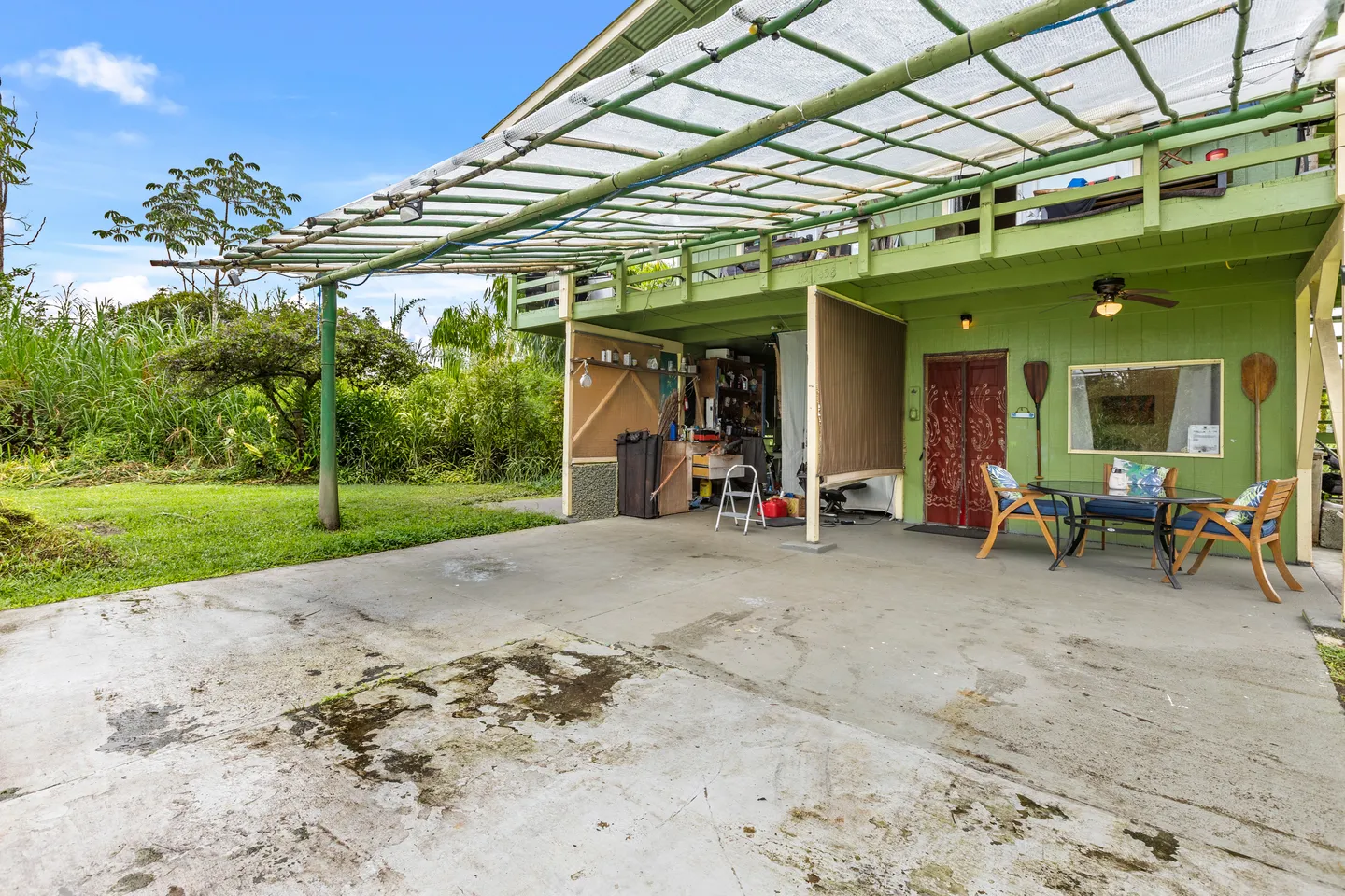 Concrete patio and carport beneath an upper deck of a green two-story house with outdoor dining seating and a lush yard.